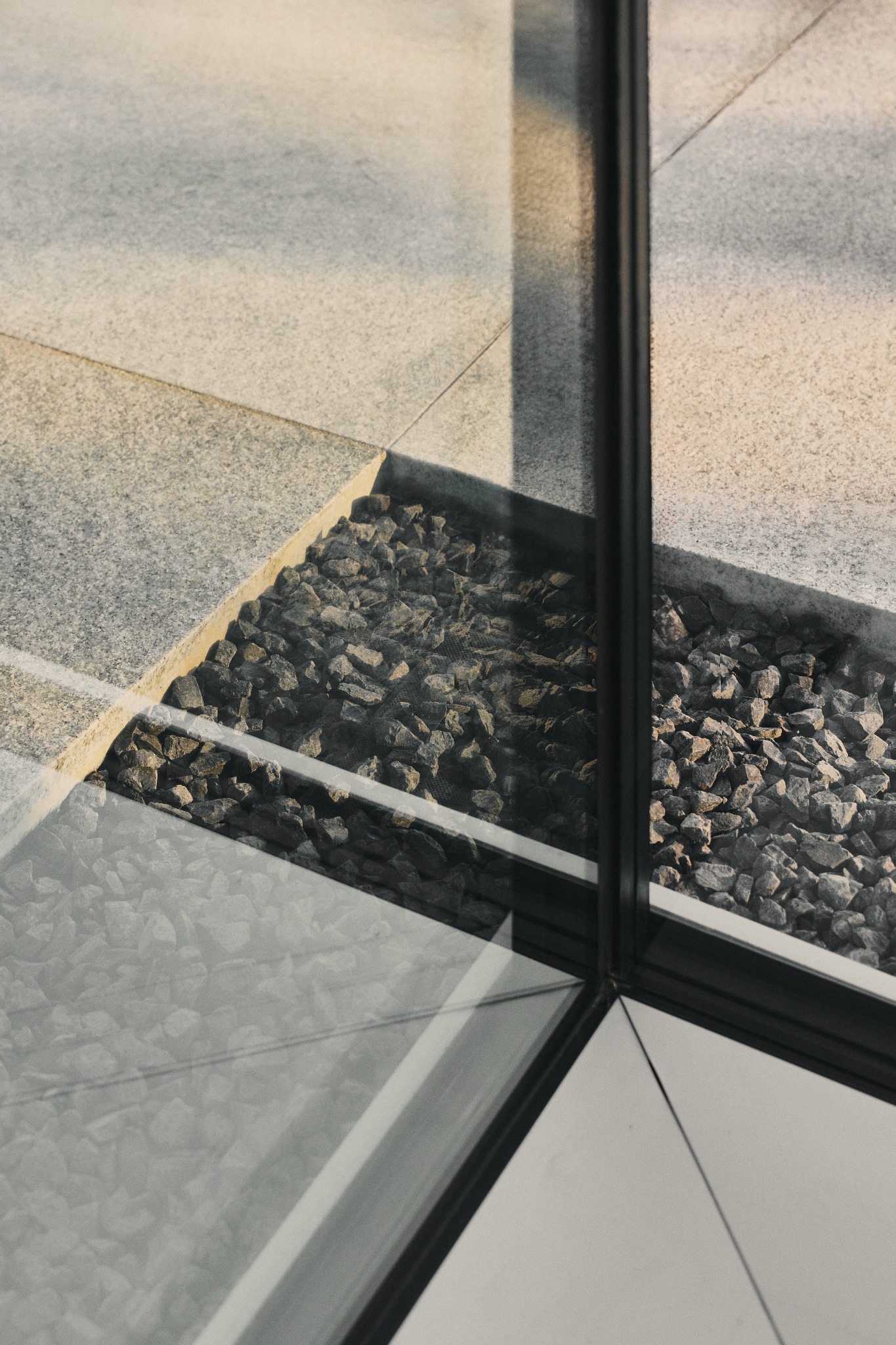 Nowoczesny detal architektoniczny: połączenie szarej kamiennej posadzki, czarnej ramy okiennej i wypełnienia z drobnych kamieni. Minimalistyczny design z wyraźnymi liniami.