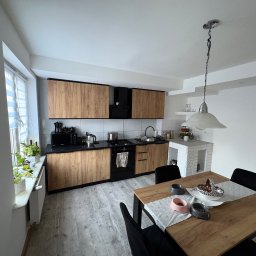 Jasna kuchnia z drewnianymi szafkami i czarnymi blatami. Na stole miski dla zwierząt. W tle widoczny biały kominek z cegły i okno z żaluzjami.