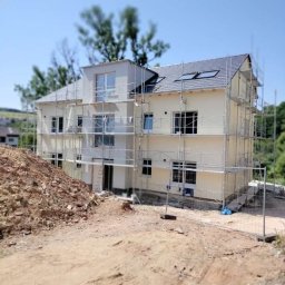 Dwupiętrowy dom w trakcie ocieplania elewacji, z widocznymi rusztowaniami i stertą ziemi na pierwszym planie. Jasna elewacja, szara dachówka, błękitne niebo w tle.