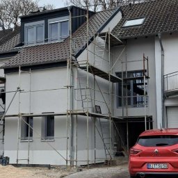 KPPS - Dom w trakcie prac elewacyjnych, widoczne rusztowania i świeżo nałożona jasna warstwa. Czerwony samochód zaparkowany obok. Ujęcie z dołu, pochmurne niebo.