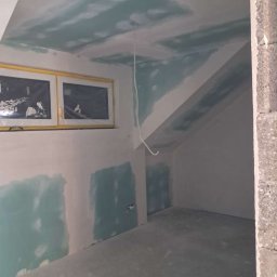 Poddasze w trakcie prac wykończeniowych: ściany i sufit pokryte częściowo gładzią, widoczne okno z żółtą ramą, przewód elektryczny zwisający z sufitu.