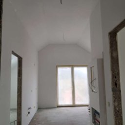 KPPS - Wnętrze po gładziowaniu ścian i sufitu, widoczne przygotowane okno do malowania, surowa podłoga i otwory drzwiowe w trakcie wykończenia.