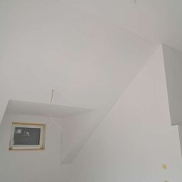 KPPS - Białe, świeżo pomalowane wnętrze poddasza z oknem zabezpieczonym folią. Widoczne skośne ściany i sufit, gładkie powierzchnie. Minimalistyczny kadr, brak dekoracji.