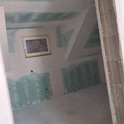 KPPS - Pokój na poddaszu w trakcie prac wykończeniowych: ściany z płyt gipsowo-kartonowych, widoczne łączenia i szpachlowanie, okno w ramie. Surowy betonowy filar w kadrze.