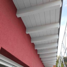 KPPS - Czerwona elewacja budynku z białym, drewnianym podbitkiem dachowym, widok z dołu pod kątem. Tekstura ściany z bliska, widoczne detale wykończenia.