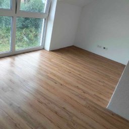 KPPS - Jasne panele podłogowe z efektem drewna w pustym pokoju z dużym oknem. Widoczne gniazdka elektryczne na białej ścianie. Ukośny sufit i ściana.