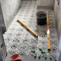 KPPS - Układanie wzorzystych płytek podłogowych w łazience, widoczne poziomice, wiadro z zaprawą i akcesoria glazurnicze. Ściany surowe, przygotowane do dalszych prac.