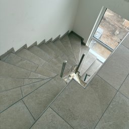 KPPS - Ukończone schody wyłożone płytkami w odcieniach szarości, widok z góry. Metalowe barierki i okno w tle. Detal: czerwony element w rogu kadru.