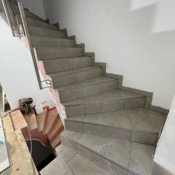 KPPS - Wykończone płytkami schody z metalową barierką, widoczne ślady prac remontowych na ścianach i podłodze. Ujęcie z dołu, ukazujące detale wykończenia.