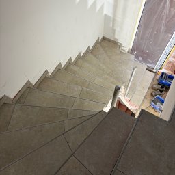 KPPS - Wykończone schody spiralne z beżowych płytek, widok z góry. Ściana w trakcie prac wykończeniowych, zabezpieczone drzwi i narzędzia glazurnicze w tle.