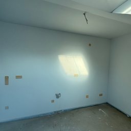 KPPS - Wnętrze pokoju po gładziowaniu ścian i sufitu, widoczne ślady po taśmach malarskich i niedoskonałości na podłodze. Świeże, białe ściany w surowym stanie.