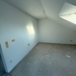 KPPS - Poddasze po nałożeniu gładzi, widoczne ślady po łatach na ścianach, surowa podłoga i okno dachowe w skosie. Minimalistyczne wnętrze w trakcie prac wykończeniowych.