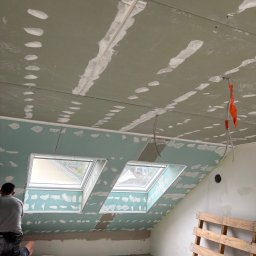 KPPS - Poddasze z oknami dachowymi w trakcie montażu płyt gipsowo-kartonowych, widoczne szpachlowane łączenia i otwory po wkrętach. Paleta oparta o ścianę, narzędzia pracy.