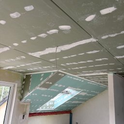 KPPS - Płyty karton-gips na skośnym suficie poddasza z oknem dachowym, widoczne łączenia płyt i miejsca po wkrętach, przygotowane do szpachlowania. Surowy stan wykończenia.