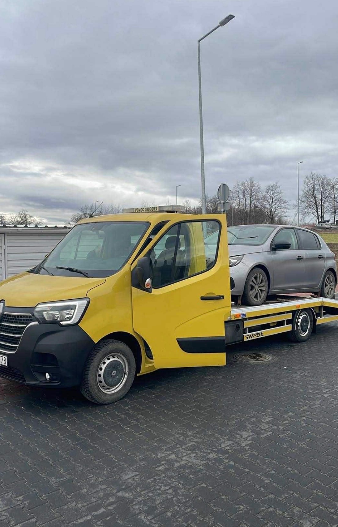 Żółta laweta Renault z otwartymi drzwiami, przewożąca srebrny samochód osobowy na platformie. Widok z boku na szarym bruku, pochmurne niebo w tle.