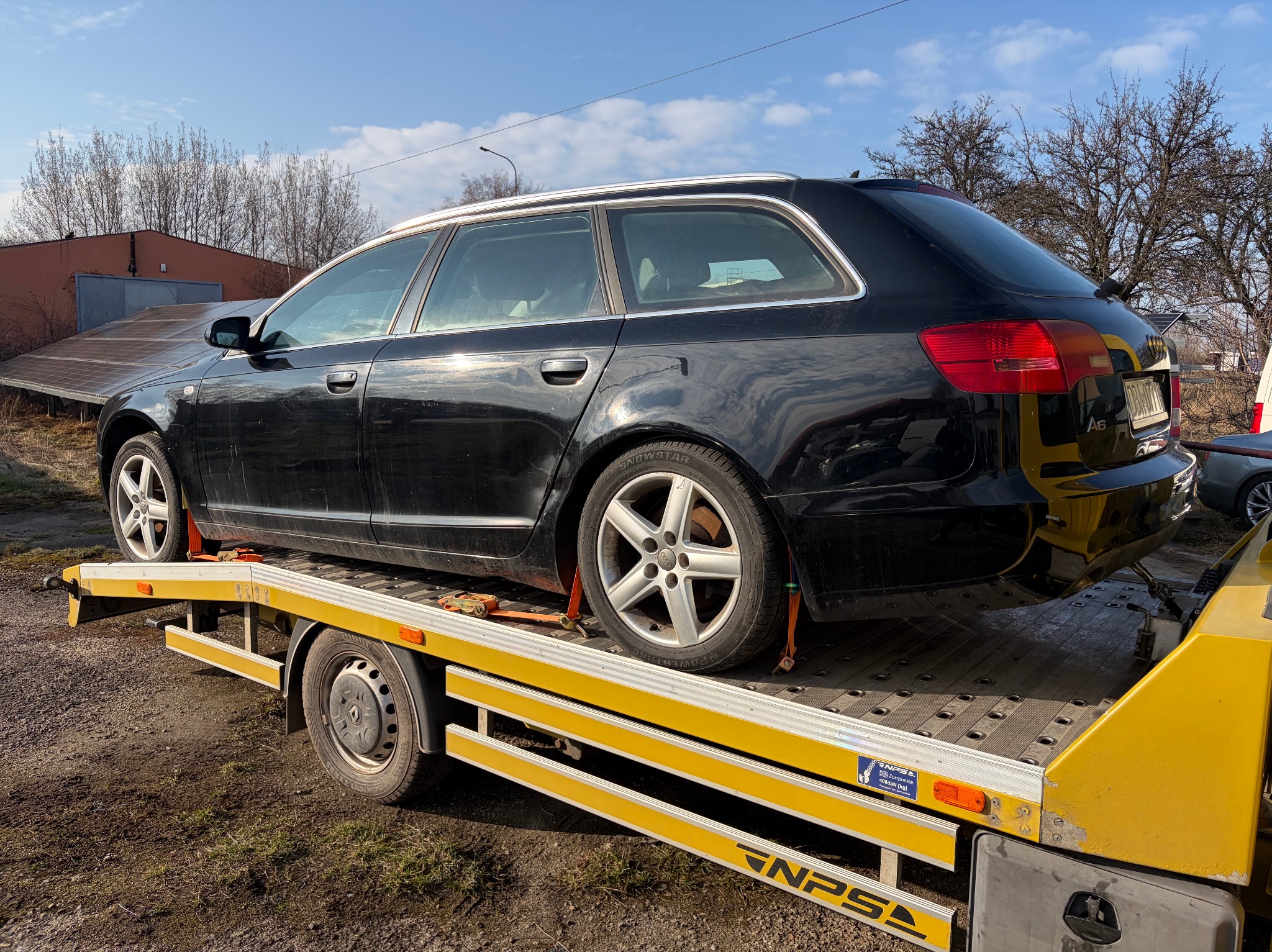 Czarna Audi A6 kombi zabezpieczona pasami na żółtej lawecie NPS, widok z tyłu, w tle budynek i drzewa bez liści, słoneczny dzień.