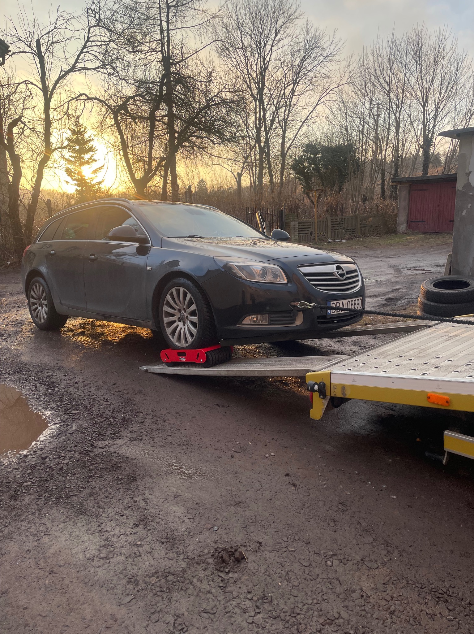 Czarny Opel Insignia na lawecie z czerwonymi wózkami pod kołami, w tle drzewa i budynek gospodarczy o zachodzie słońca. Rejestracja widoczna.