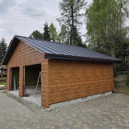 Trans-Blach2 - Nowoczesny garaż dwustanowiskowy z blachy trapezowej w kolorze antracytowym, wykończony drewnopodobną elewacją. Widoczne elementy konstrukcyjne i otoczenie.