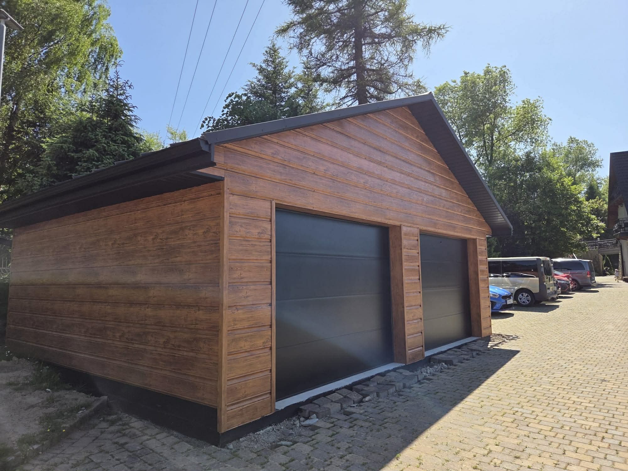 Drewniany garaż dwustanowiskowy z czarnymi bramami segmentowymi, wykończony poziomymi deskami. Widoczne czarne rynny i kostka brukowa przed budynkiem.
