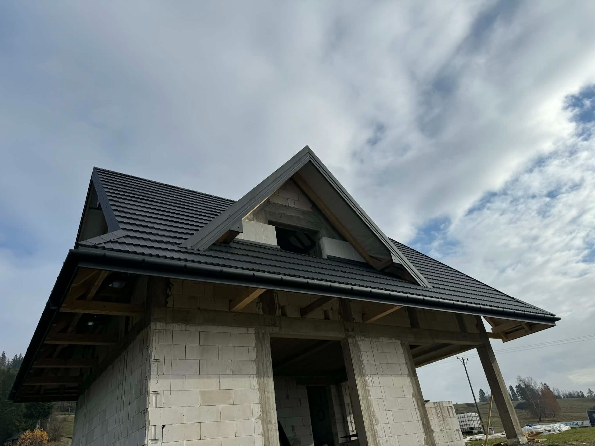 Dom w stanie surowym z czarnym dachem i rynnami, widok z dołu na tle pochmurnego nieba. Widoczne elementy konstrukcyjne i materiały budowlane.