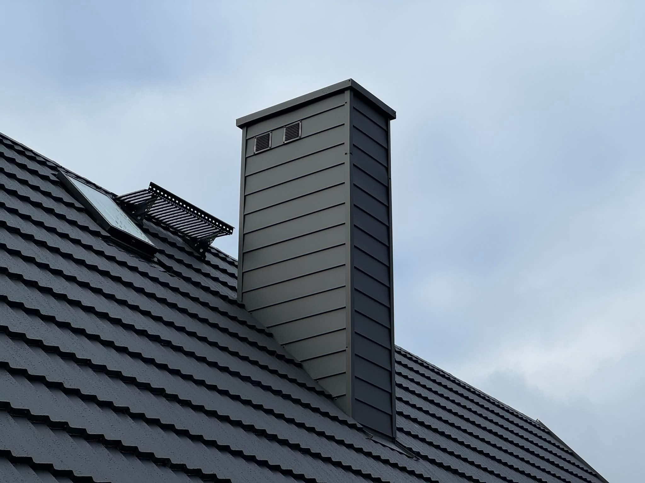 Szary komin z blachy na dachu z dachówką ceramiczną, widok z dołu. Na dachu okno połaciowe i kolektor słoneczny. Pochmurne niebo w tle.