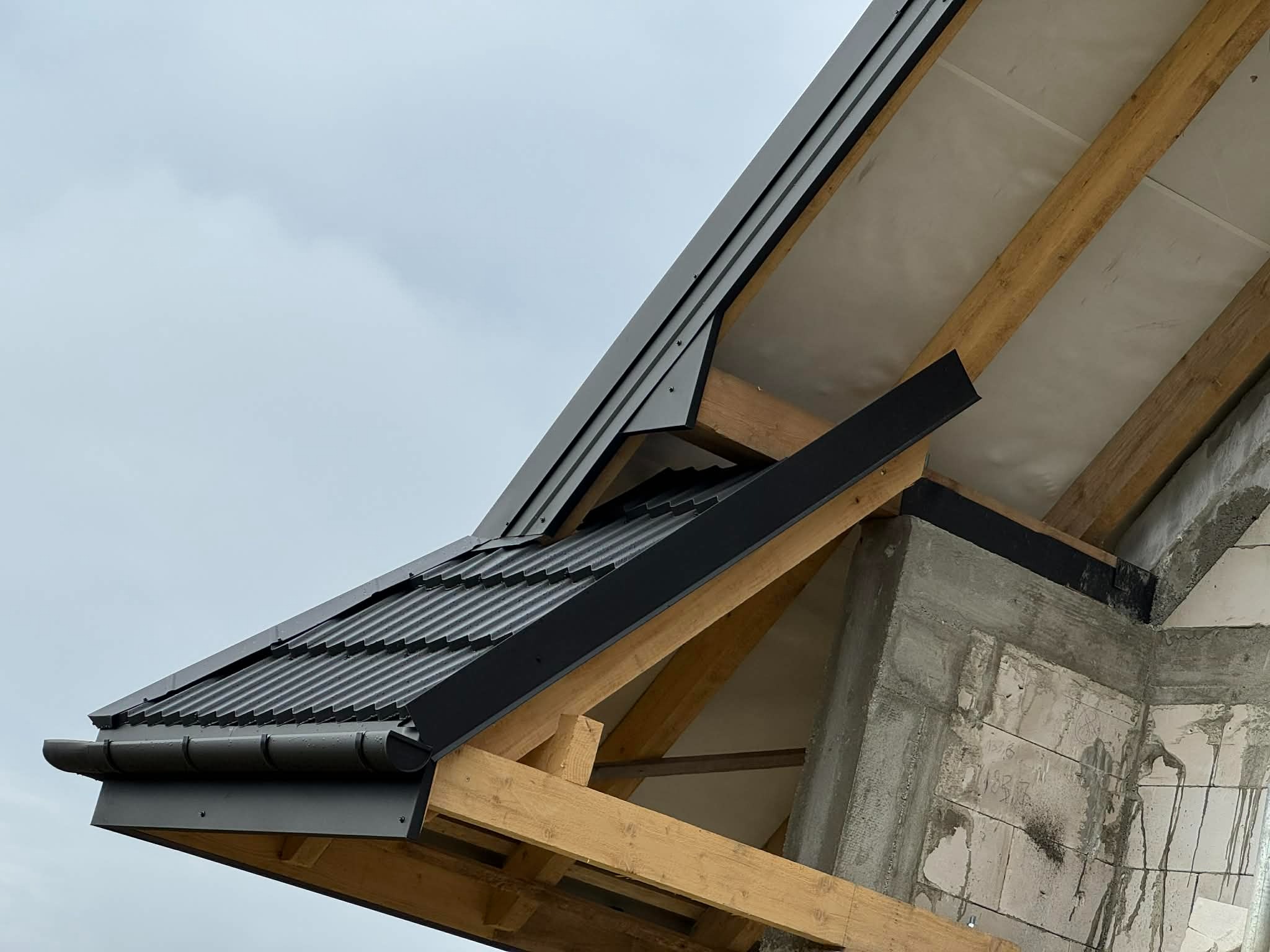 Fragment dachu w trakcie budowy, widoczne drewniane krokwie i czarne elementy wykończenia dachu. Ujęcie z dołu, pochmurne niebo w tle.