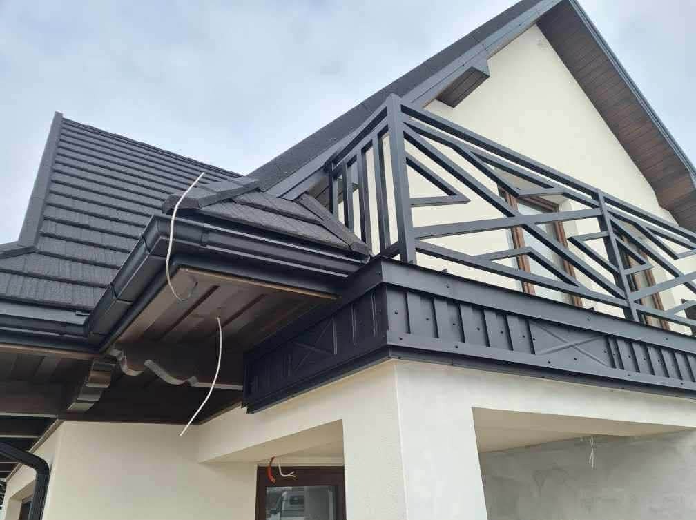 Nowoczesny dom z ciemną dachówką, czarnymi rynnami i balustradą balkonu. Widoczne detale obróbek blacharskich i podbitki dachowej. Ujęcie z dołu, pochmurne niebo.