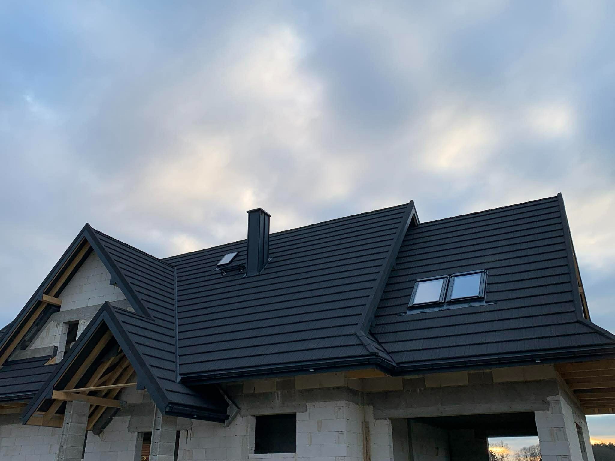 Dom w trakcie budowy z czarnym dachem z dachówki modułowej, widoczne okna dachowe i komin. Budynek z białego bloczka, częściowo otynkowany. Pochmurne niebo w tle.