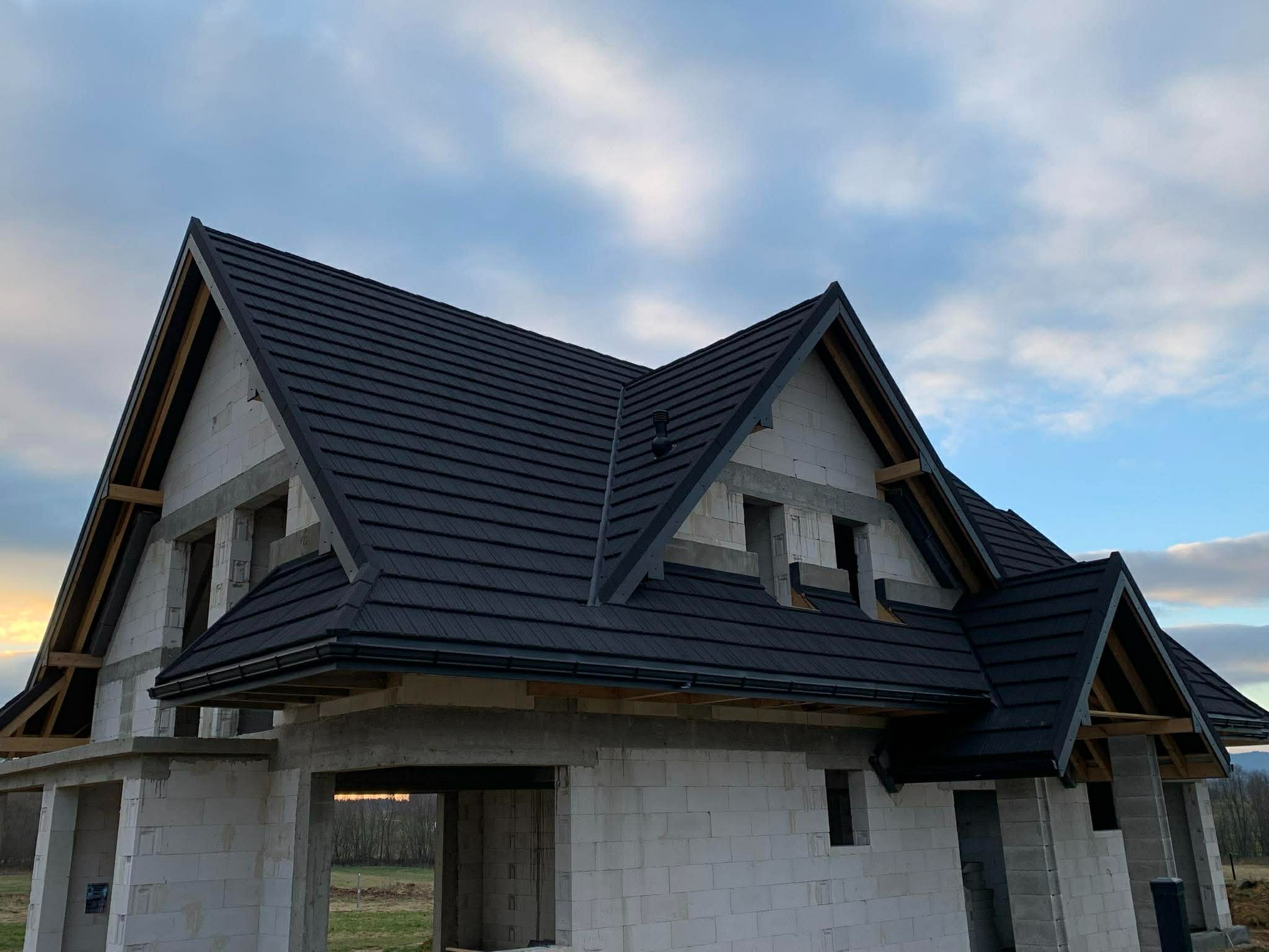 Czarny dach z blachy na surowym budynku w Rabce-Zdroju. Widoczne rynny i obróbki blacharskie. Dachówka modułowa na tle pochmurnego nieba.