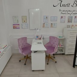 ANETT BEAUTY ANETA KRĘCICHWOST - Jasne wnętrze salonu kosmetycznego z dwoma fotelami i stanowiskiem do manicure, ozdobione certyfikatami i dekoracjami. Widoczny parawan.