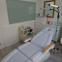 ANETT BEAUTY ANETA KRĘCICHWOST - Jasne wnętrze gabinetu kosmetycznego z białym fotelem zabiegowym, sprzętem kosmetycznym i plakatem anatomicznym. Widok na drzwi z grafiką i okno.