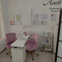 ANETT BEAUTY ANETA KRĘCICHWOST - Jasne wnętrze salonu kosmetycznego z fotelem i stolikiem do manicure, ozdobione certyfikatami i dekoracyjnym parawanem.