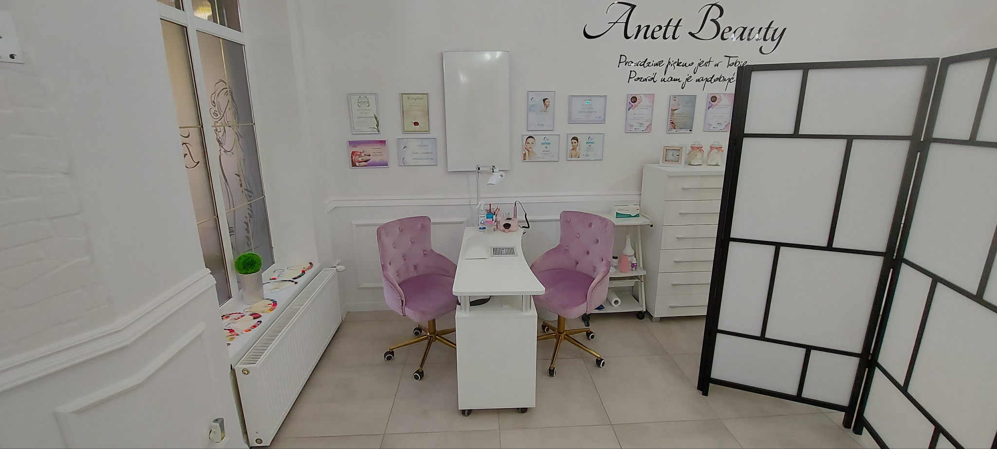 Jasne wnętrze salonu kosmetycznego z dwoma fotelami i stanowiskiem do manicure, ozdobione certyfikatami i dekoracjami. Widoczny parawan.