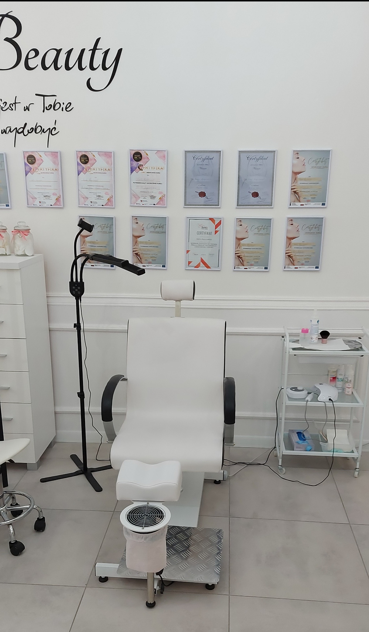 Jasne wnętrze salonu z fotelem kosmetycznym, lampą i wózkiem z akcesoriami. Na ścianie wiszą certyfikaty. Minimalistyczny wystrój, czyste linie.