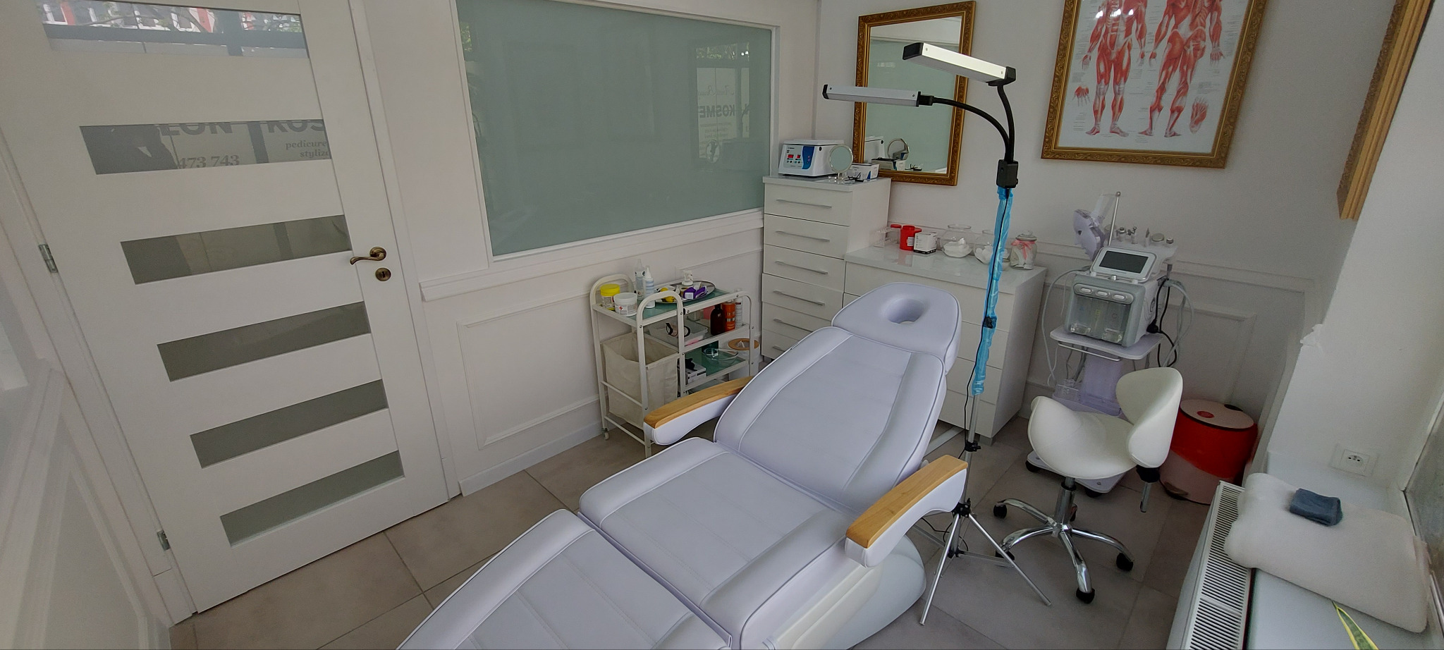 Jasne wnętrze gabinetu kosmetycznego z białym fotelem zabiegowym, sprzętem kosmetycznym i plakatem anatomicznym. Widok na drzwi z grafiką i okno.