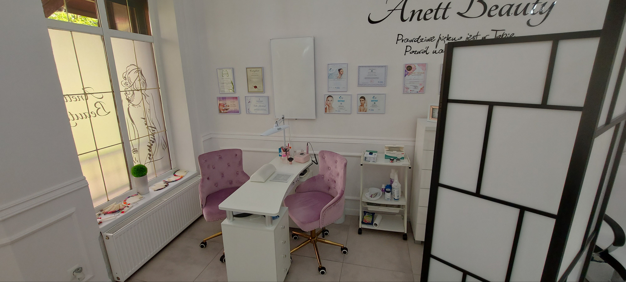 Jasne wnętrze salonu kosmetycznego z fotelem i stolikiem do manicure, ozdobione certyfikatami i dekoracyjnym parawanem.
