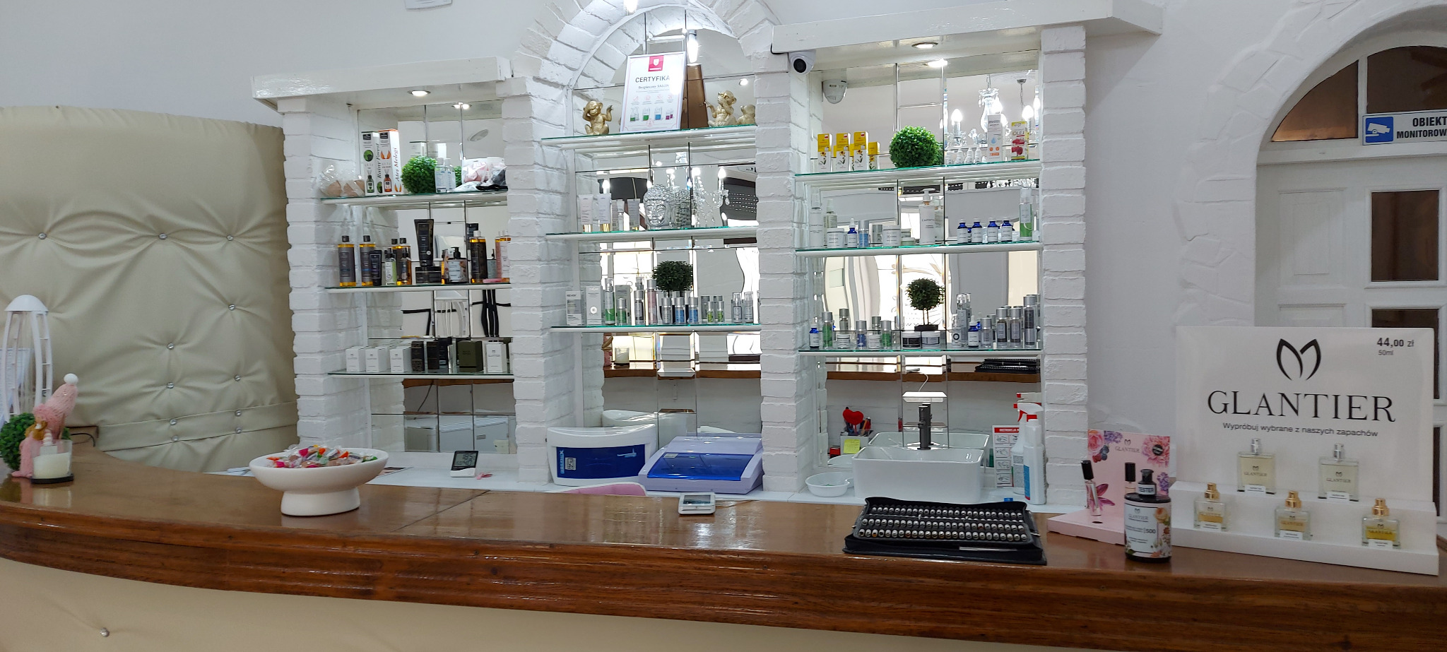 Jasne wnętrze salonu kosmetycznego z ekspozycją produktów na półkach i ladą recepcyjną z testerami perfum Glantier. Widoczny certyfikat i dekoracje.