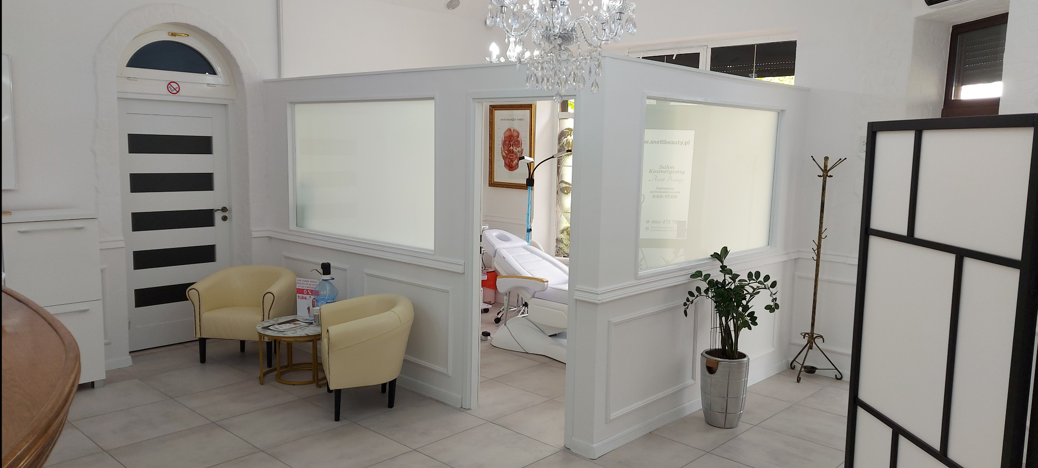 Jasne wnętrze salonu kosmetycznego z kremowymi fotelami, kryształowym żyrandolem i kozetką zabiegową w tle. Widoczny plakat z ofertą salonu.