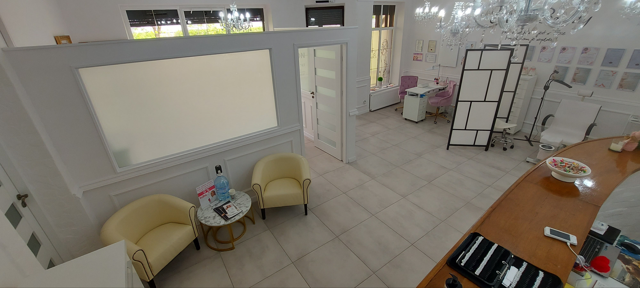 Jasne wnętrze salonu kosmetycznego z recepcją, fotelami dla klientów i stanowiskiem do manicure. Widoczne detale wystroju, takie jak żyrandole i dekoracje ścienne.