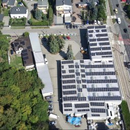 Instal PV - Widok z drona na budynek z panelami słonecznymi na dachu, otoczony zielenią i ulicą z ruchem. Panele ułożone w rzędach, widoczne budynki mieszkalne w tle.
