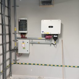 Instal PV - Zakończona instalacja fotowoltaiczna wewnątrz budynku: inwerter Huawei, rozdzielnica, kable w korytach, uziemienie. Widoczna taśma ostrzegawcza i szczotka.