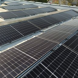 Instal PV - Rząd paneli fotowoltaicznych na płaskim dachu, widok z góry, szare mocowania, panele w czarnych ramach, w tle drzewa i budynek.