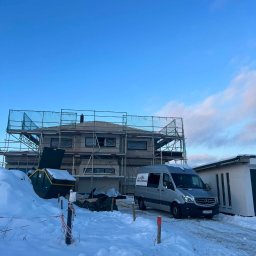 Wasyl Moroc - Dom w trakcie budowy, otoczony rusztowaniami i siatką zabezpieczającą. Na pierwszym planie zaspy śnieżne, kontener na odpady i samochód dostawczy z logo firmy.