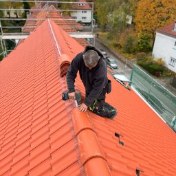 Wasyl Moroc - Monter w czarnej bluzie, klęcząc na pomarańczowym dachu z dachówki ceramicznej, przykręca element wzdłuż kalenicy. W tle drzewa i budynki.