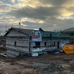 Wasyl Moroc - Budowa domu modułowego w trakcie: szare ściany, rusztowania, dach z zielonej dachówki. Na dachu widoczny pracownik. Żółty kontener na gruz. Pochmurne niebo.