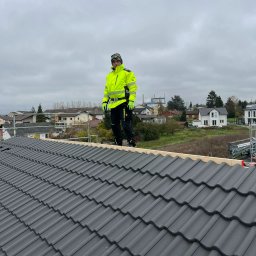 Wasyl Moroc - Dekarz w jaskrawej kurtce na dachu z szarej dachówki, w tle zabudowa jednorodzinna i pochmurne niebo. Widoczny fragment rusztowania.