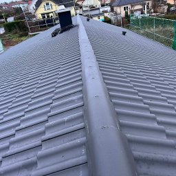 Wasyl Moroc - Szary dach z dachówki ceramicznej, widok z góry, z kominem i zabezpieczeniami na krawędzi. Widoczne domy w tle. Dach po renowacji lub nowo położony.