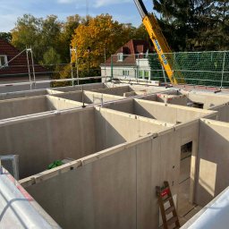 Wasyl Moroc - Wznoszenie domu modułowego: widok z góry na konstrukcję ścian z otworami okiennymi i drzwiowymi, z dźwigiem w tle i jesiennym krajobrazem.