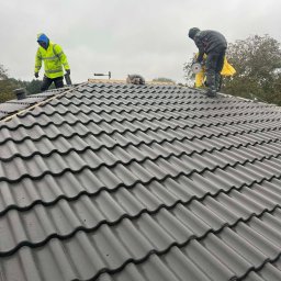 Wasyl Moroc - Montaż ciemnoszarej dachówki ceramicznej na dachu przez dwóch dekarzy, jeden z nich używa szlifierki kątowej. Widoczne narzędzia i elementy konstrukcji dachu.