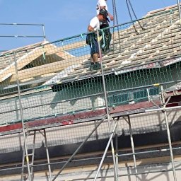 Wasyl Moroc - Praca na dachu z rusztowania: dekarze montują drewnianą konstrukcję dachu, zabezpieczeni siatką. Widoczne elementy domu w tle i łańcuch podtrzymujący materiały.